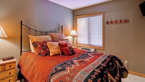 Quarto com cama, mesa de cabeceira, abajur, janela com persianas e um cabideiro na parede.