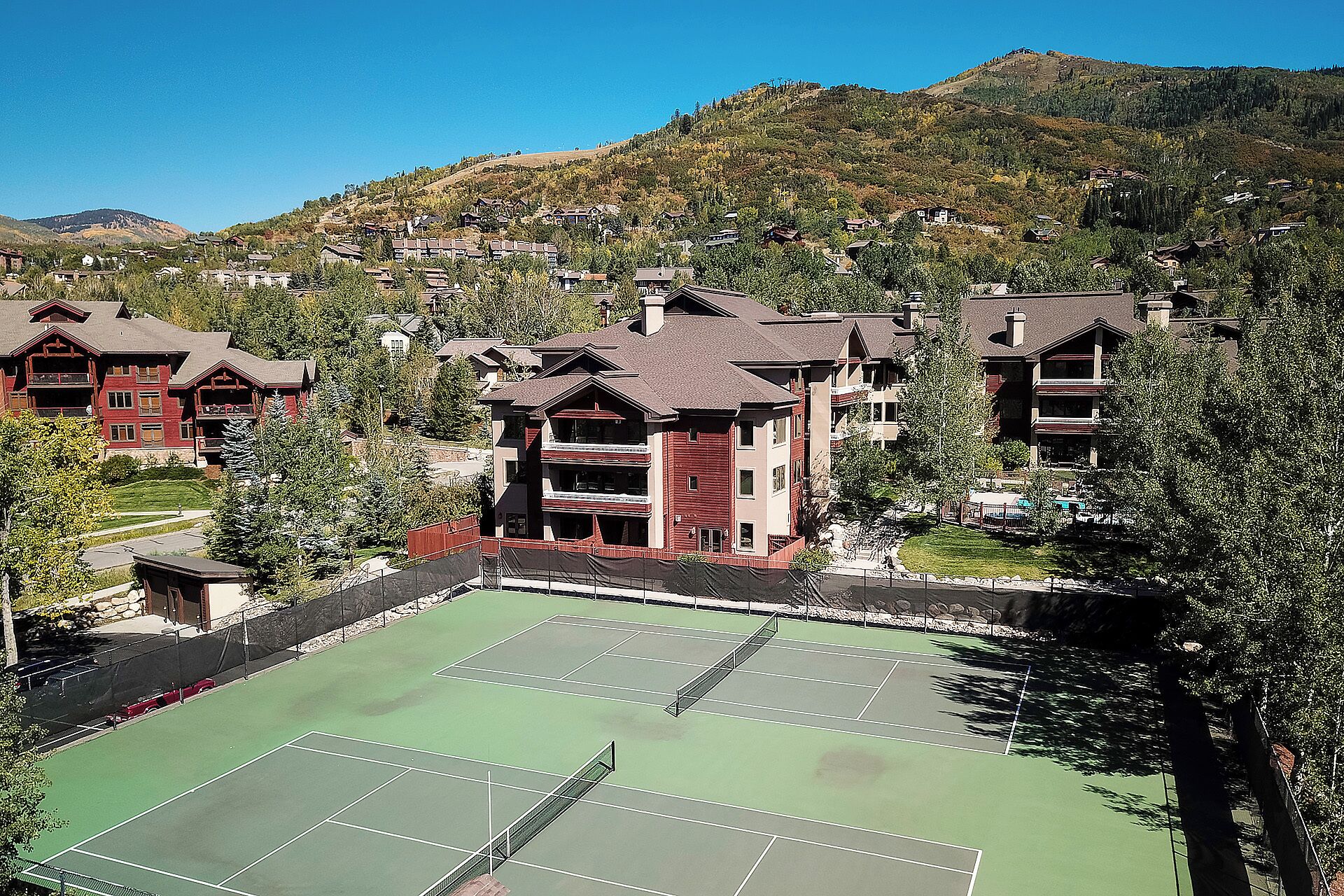 A tennis court surrounded by multi-story buildings with balconies, set against a backdrop of hills and trees.