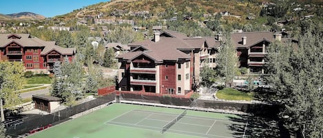Un court de tennis entouré d’immeubles à plusieurs étages dotés de balcons, avec en arrière-plan des collines et des arbres.