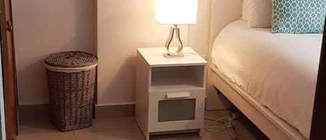 Un dormitorio con cama, una mesita de noche con lámpara, una cesta de mimbre y un cuadro en la pared.