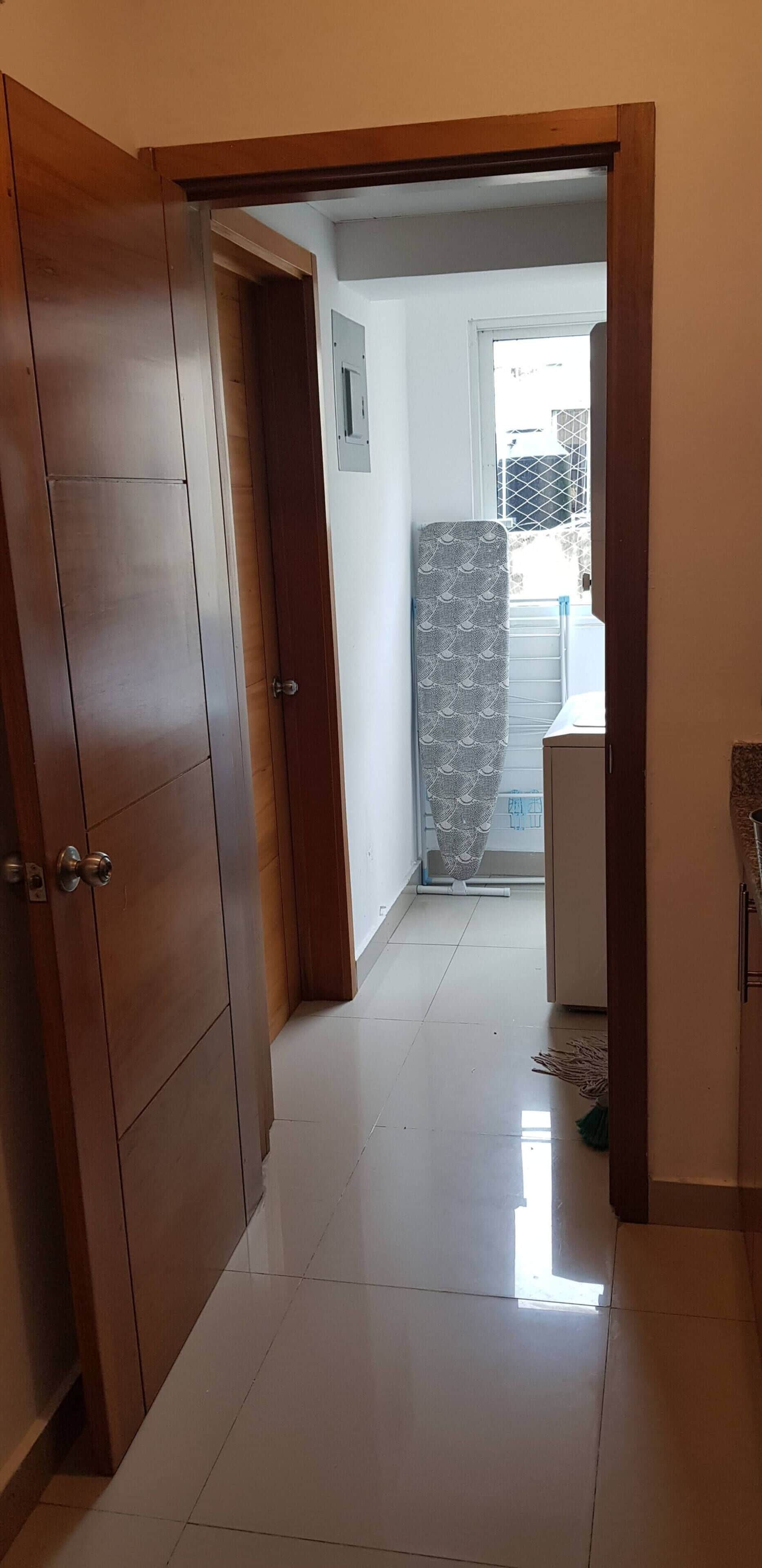 A hallway with a wooden door, a tiled floor, and a view into a room with a bed and a window.