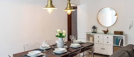 Un coin repas avec une table en bois dressée pour quatre personnes, des chaises transparentes et deux lampes suspendues.
