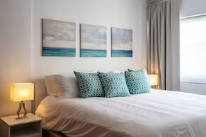 A hotel room with a bed, two bedside lamps, a window with curtains, and three wall-mounted paintings. - 2br Kingbed Kids Play Area No Surgery (Santo Domingo)