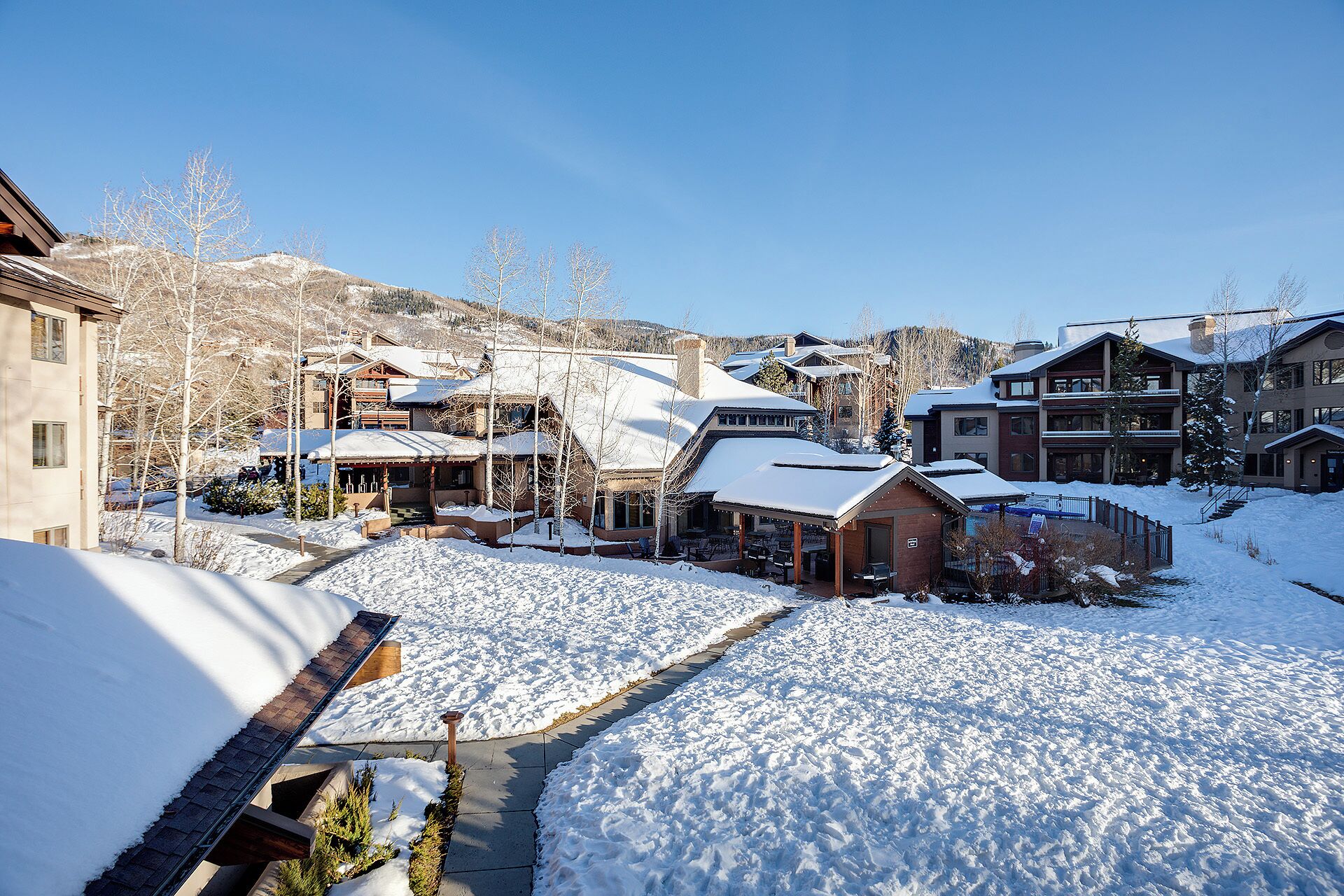 A snow-covered residential area with multiple houses, a clear sky, and a hilly background.
