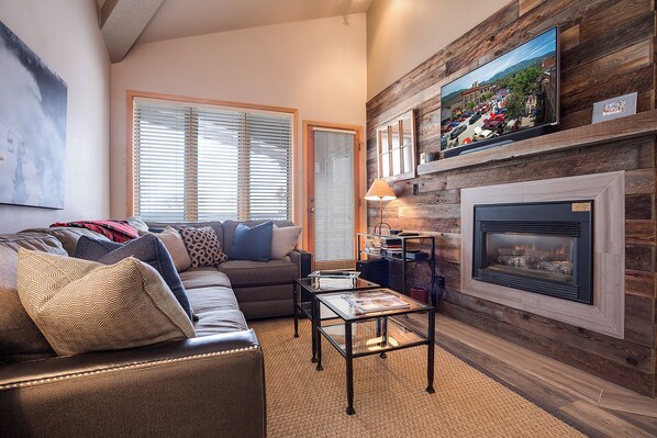A cozy living room with a fireplace, a sofa, and a television mounted on a wooden wall. - TR1302 | Nicely Updated Condo | Ski Lockers | A/C | 10 Hot Tubs (Steamboat Springs)