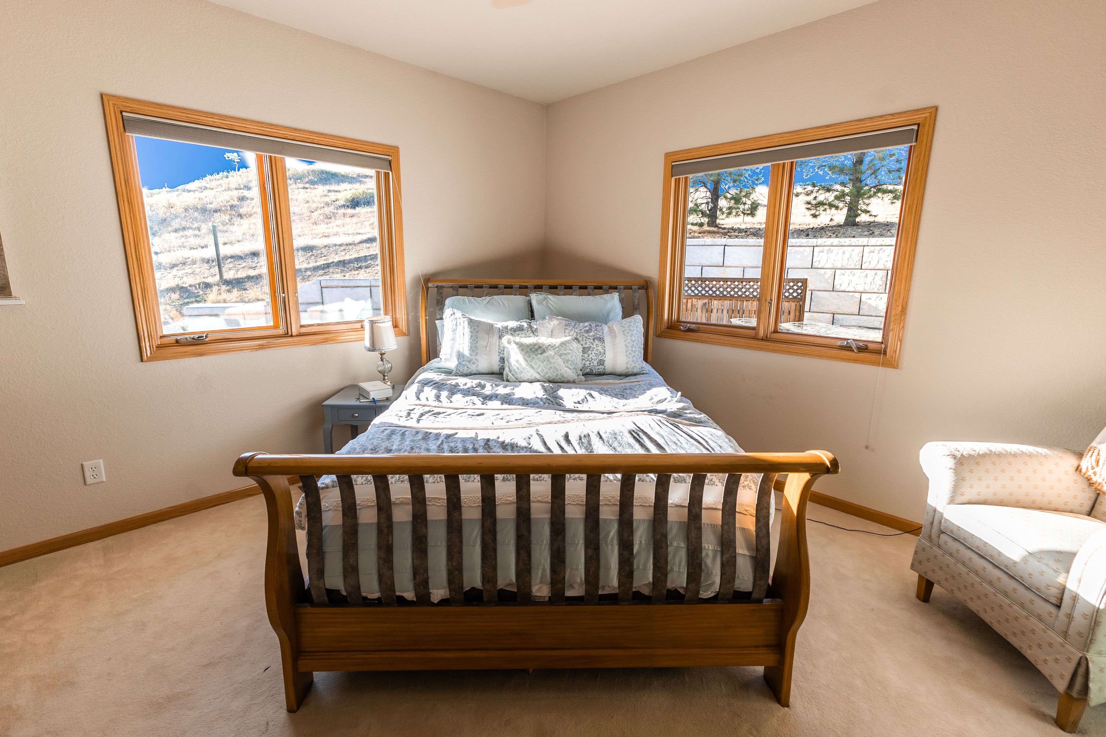 A bedroom with a wooden bed, two windows with views, a nightstand, and a chair.