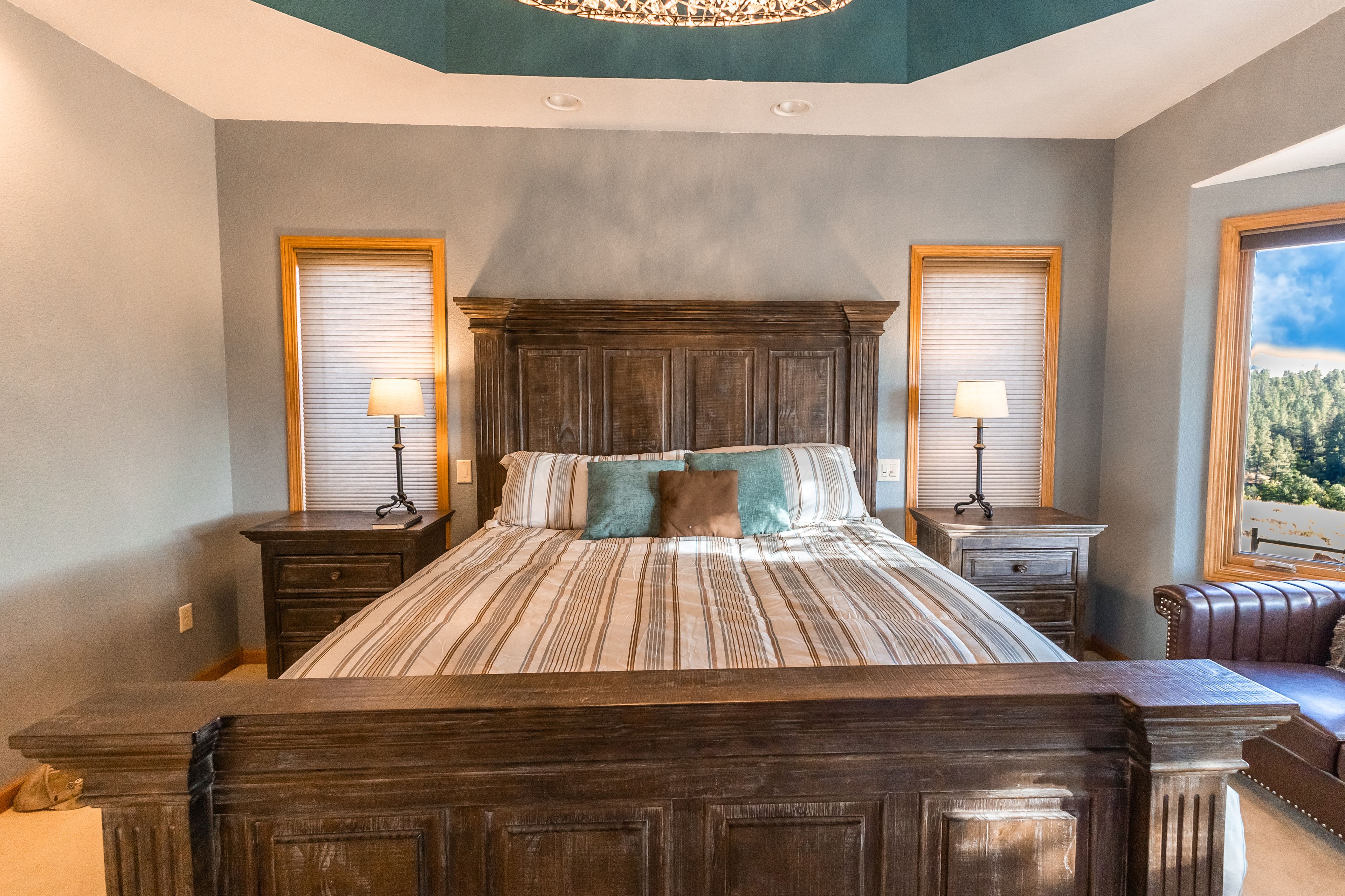 A bedroom with a large bed, two bedside tables with lamps, a headboard, and a view of a forest through a window.