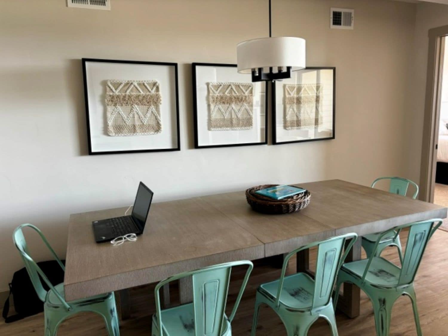 A dining area with a long table, four chairs, a laptop, and framed artwork on the wall.