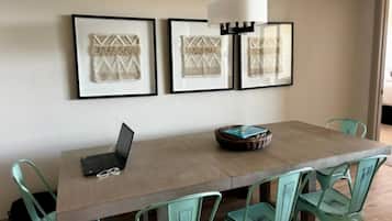 A dining area with a long table, four chairs, a laptop, and framed artwork on the wall.