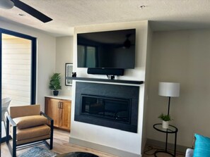 A living room with a fireplace, a television mounted above it, a chair, a small table with a lamp, and a potted plant. - Bear Lake Family Escape - Sleeps 16 - Beach access, walk to Restaurants & Fun! (Garden City)