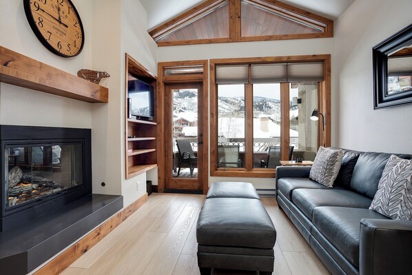 A living room with a fireplace, a sofa, a television on a wooden shelf, and a view of snowy mountains through large windows. - TR1307 | Recently Updated | Gorgeous and Unique Space | A/C | Hot Tub (Steamboat Springs)