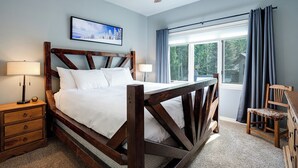 Un dormitorio con estructura de cama de madera, mesita de noche, lámpara, ventana con cortinas y ventilador de techo.