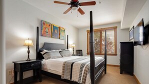 Ein Schlafzimmer mit einem Himmelbett, zwei Nachttischen inklusive Lampen, einem Deckenventilator, einem Fenster mit Jalousien und einem an der Wand befestigten Fernseher.