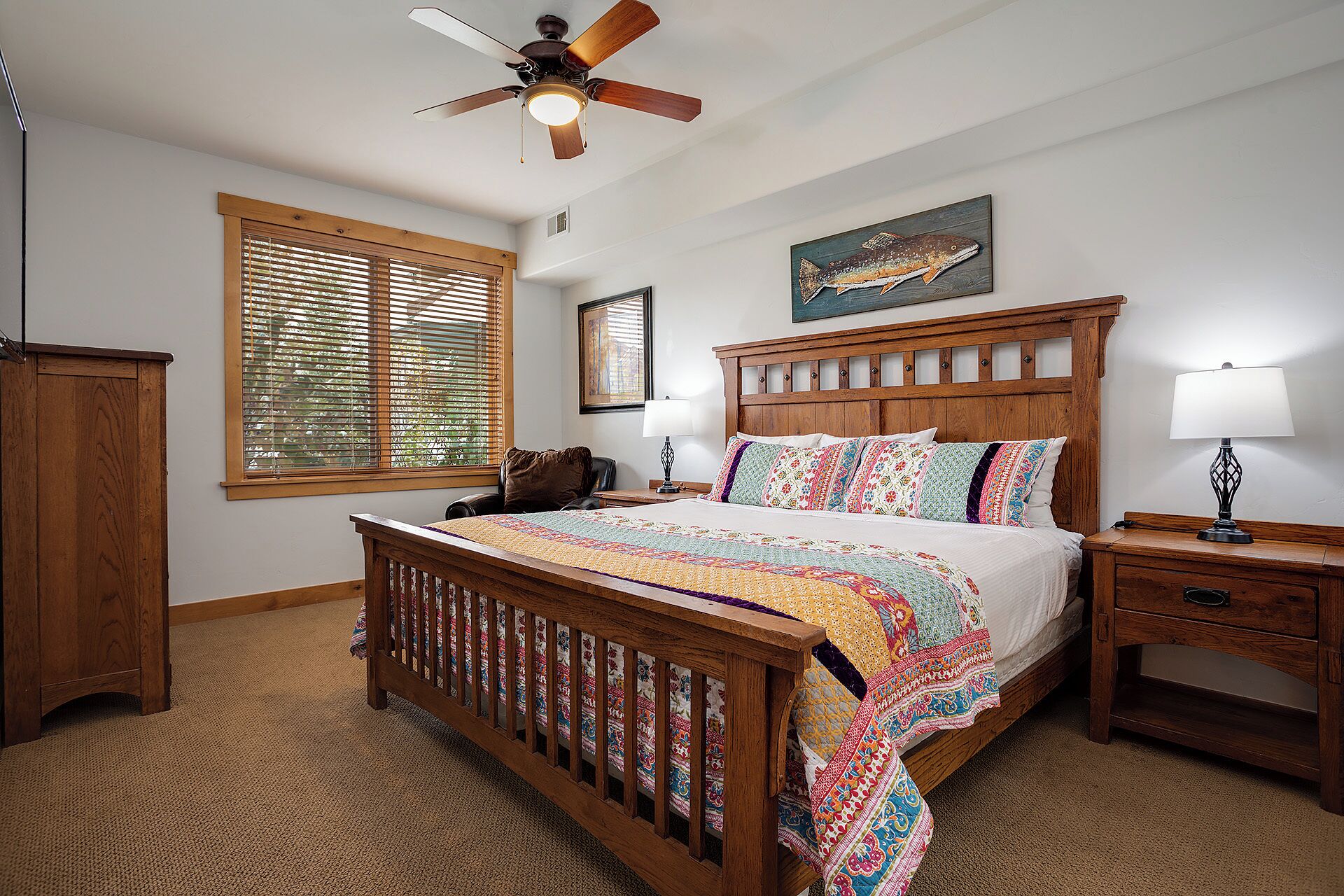 A bedroom with a wooden bed, a ceiling fan, a window with blinds, a nightstand, and a lamp.