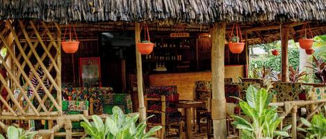 A rustic thatched-roof structure with a wooden lattice wall, a central wooden table, and potted plants.