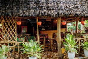 A rustic thatched-roof structure with a wooden lattice wall, a central wooden table, and potted plants.