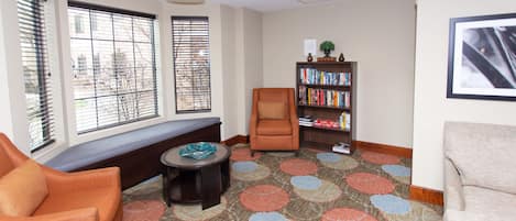 A room with a bench, two armchairs, a small table, a bookshelf, and large windows with blinds.