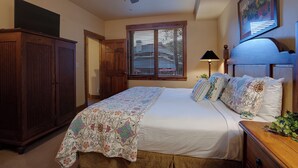Quarto com cama, ventoinha no teto, janela com persianas, mesa de cabeceira e guarda-roupa de madeira.