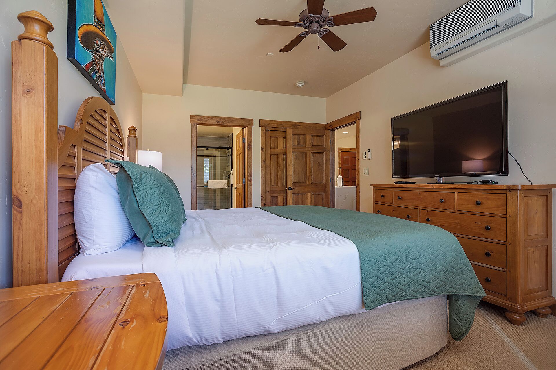 A bedroom with a bed, wooden headboard, a television, a ceiling fan, and a door leading to another room.