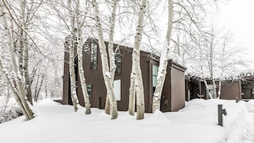 A snow-covered wooden cabin with a window and a door, surrounded by trees.