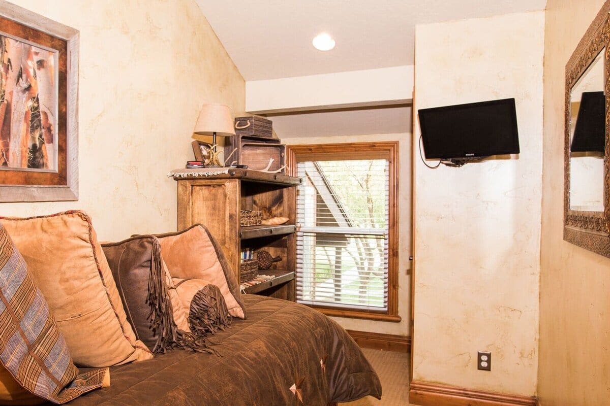Una habitación con un sofá marrón, un armario de madera con varios objetos, un televisor montado en la pared y una ventana con persianas.