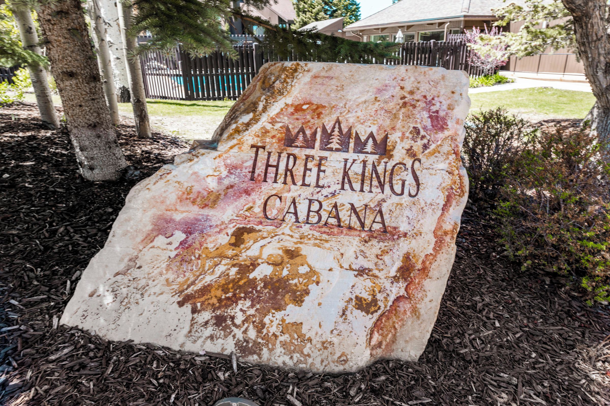 A weathered sign with the text 'THREE KINGS CABANA' in a rustic font, surrounded by mulch and trees.