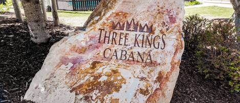 A weathered sign with the text 'THREE KINGS CABANA' in a rustic font, surrounded by mulch and trees.