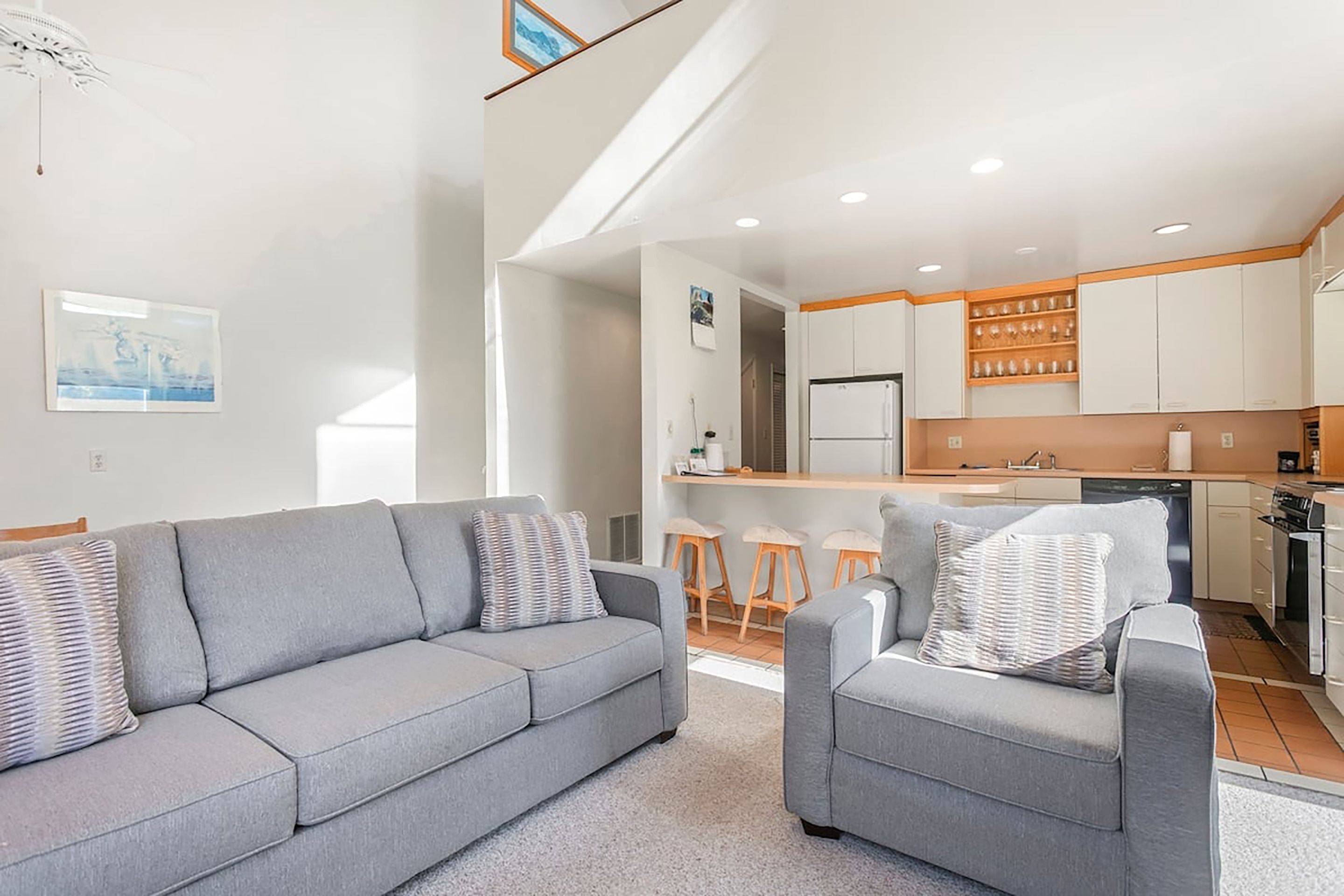 A modern living room with a grey sofa and armchairs, a kitchen with white cabinets and a wooden countertop, and a small dining area with stools.