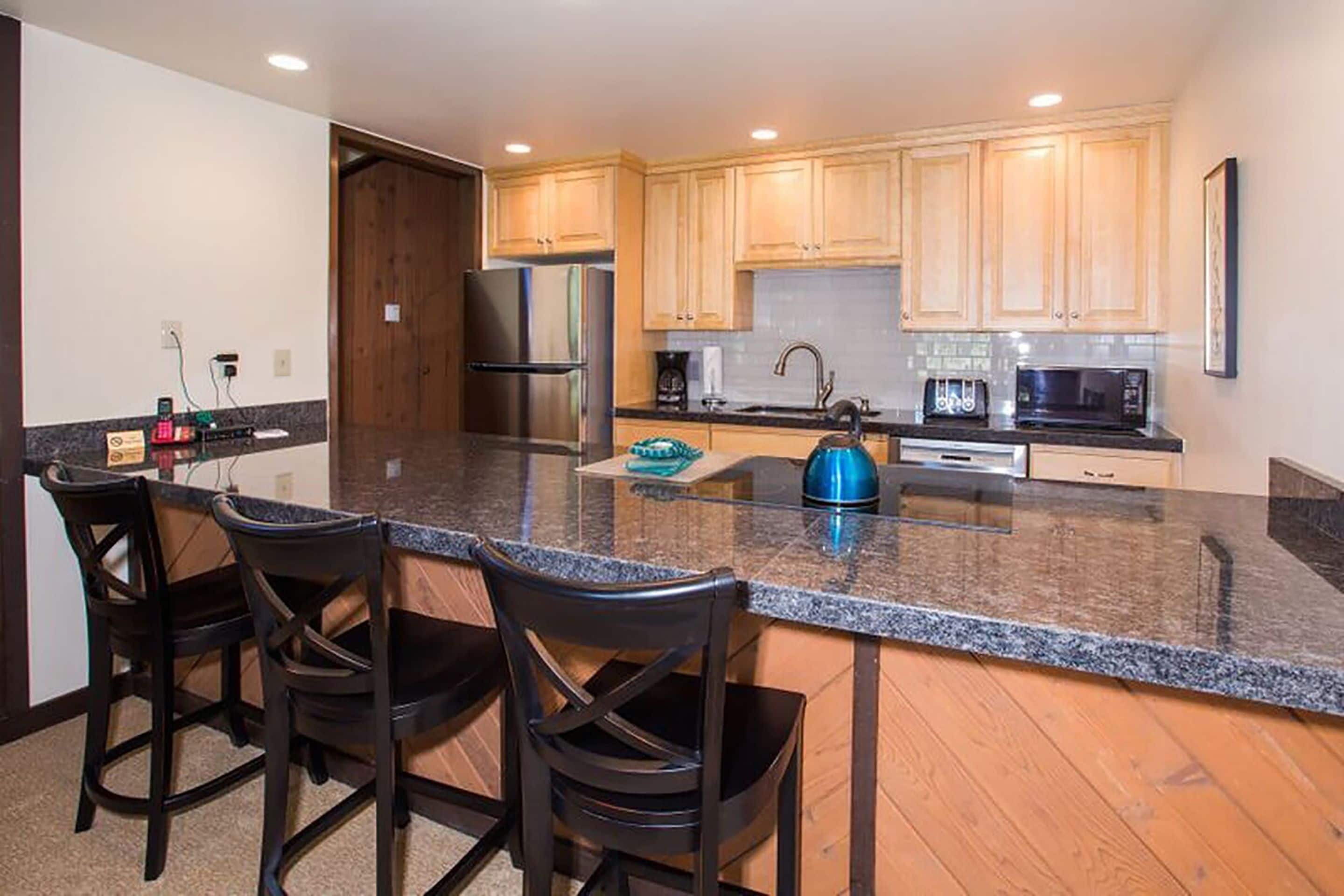 A kitchen with a granite countertop, stainless steel appliances, and wooden cabinets. There are three bar stools and a kettle on the counter.