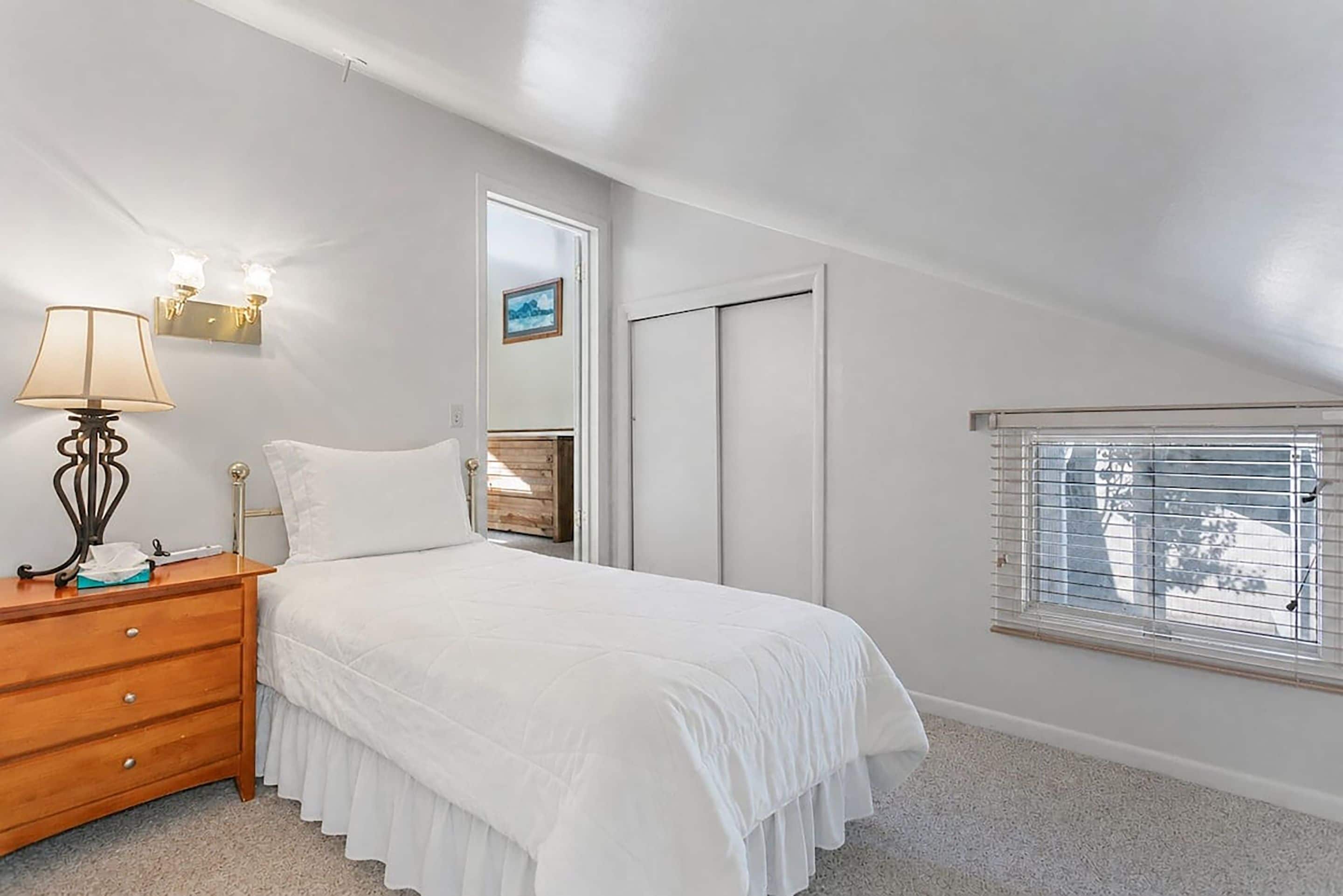 A bedroom with a bed, a nightstand, a lamp, a mirror, a wardrobe, and a window with blinds.