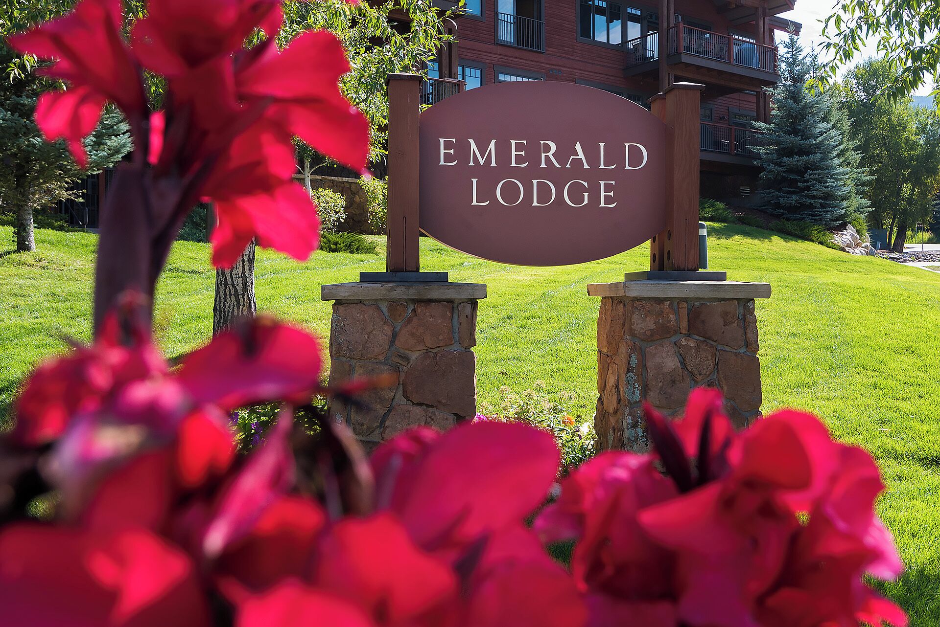 Placa para Emerald Lodge com base de pedra e poste de madeira, ao fundo um edifício e vegetação.
