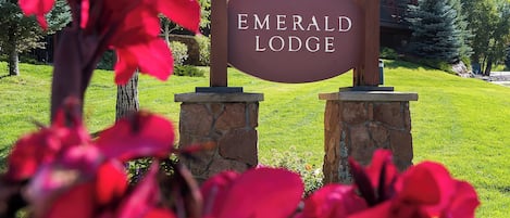 Naamplaatje voor Emerald Lodge, met een stenen voetstuk en houten paal, voor een gebouw en groen.