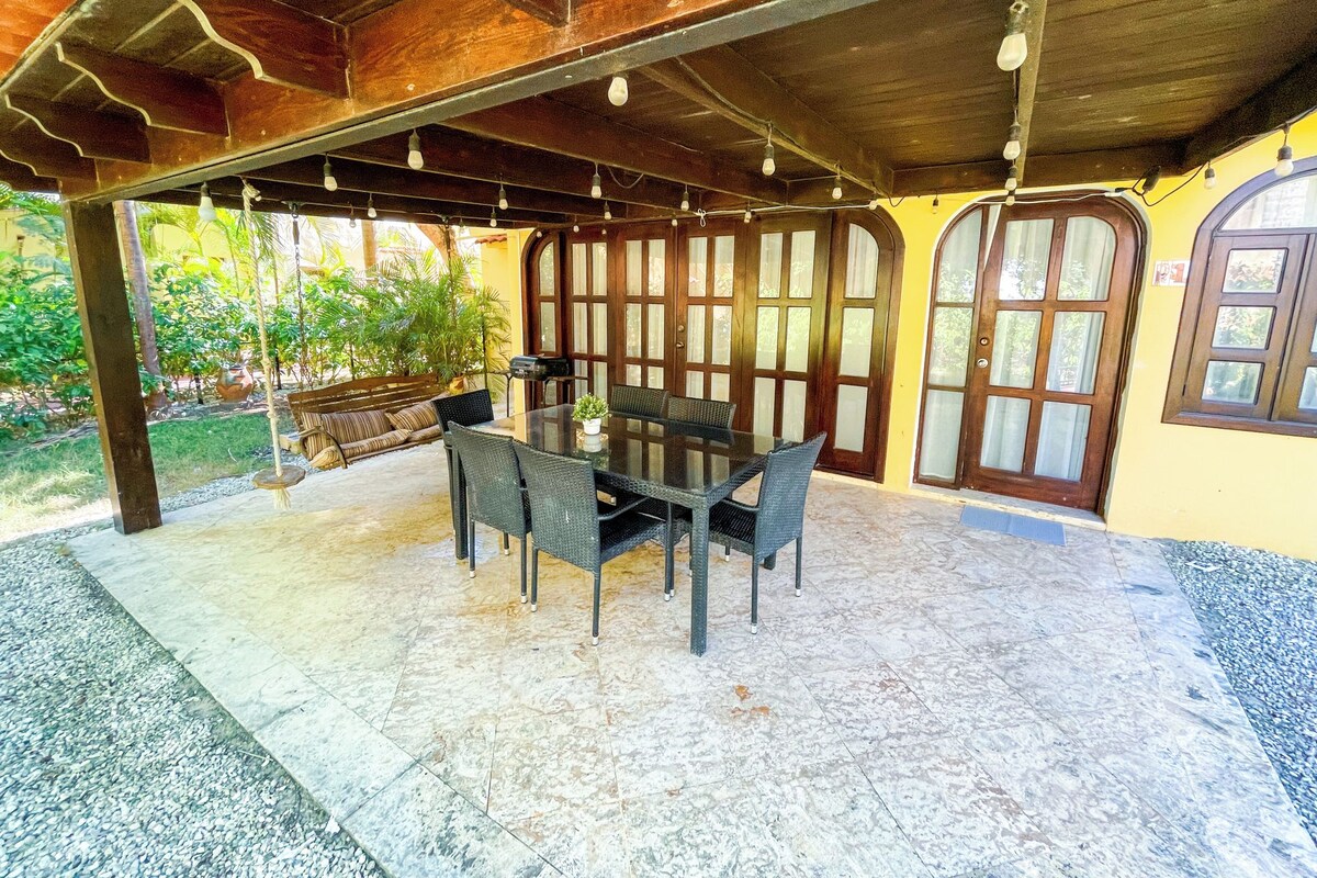 Um pátio externo com mesa e cadeiras de vidro, cercado por portas de madeira e piso de azulejos.