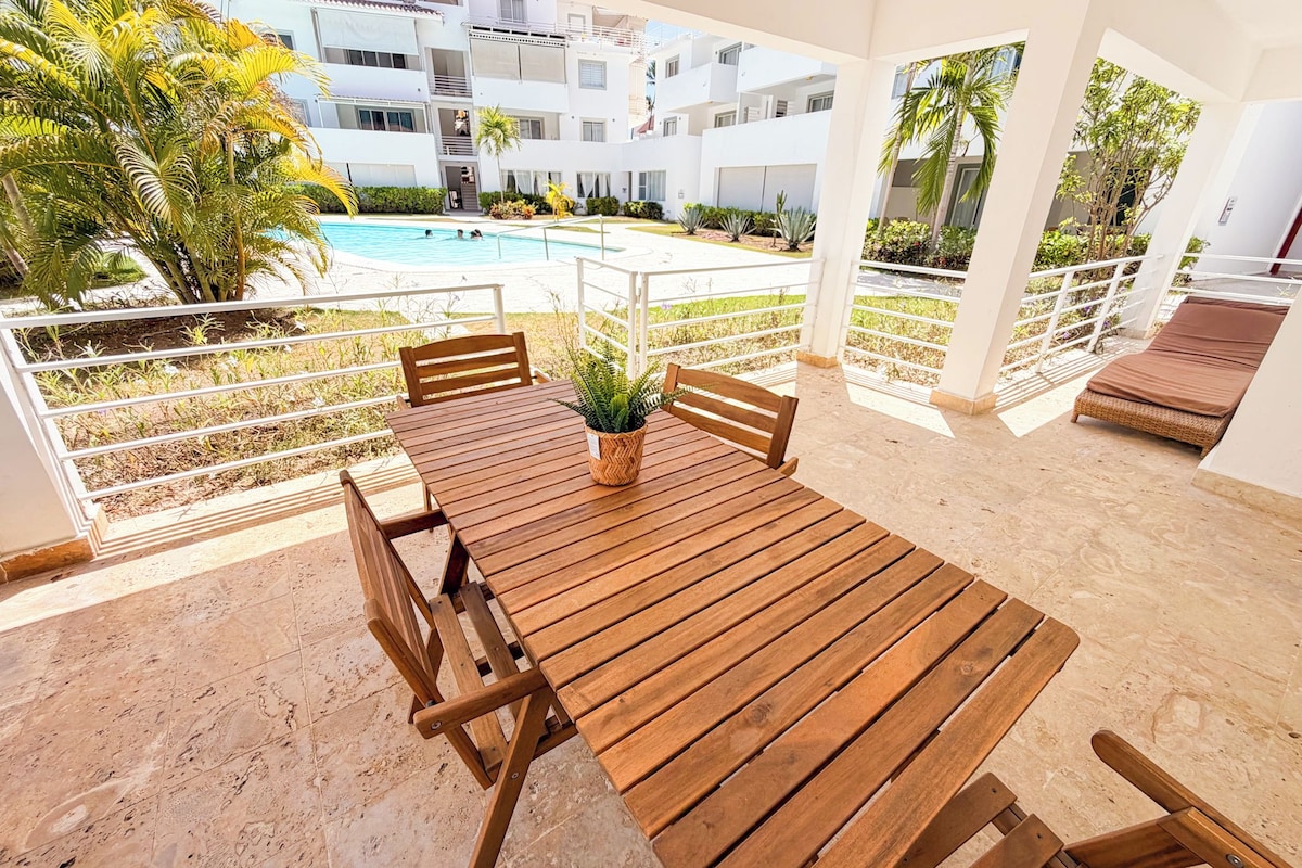 Mesa de madeira com cadeiras e um vaso de planta numa varanda com vista para uma piscina.