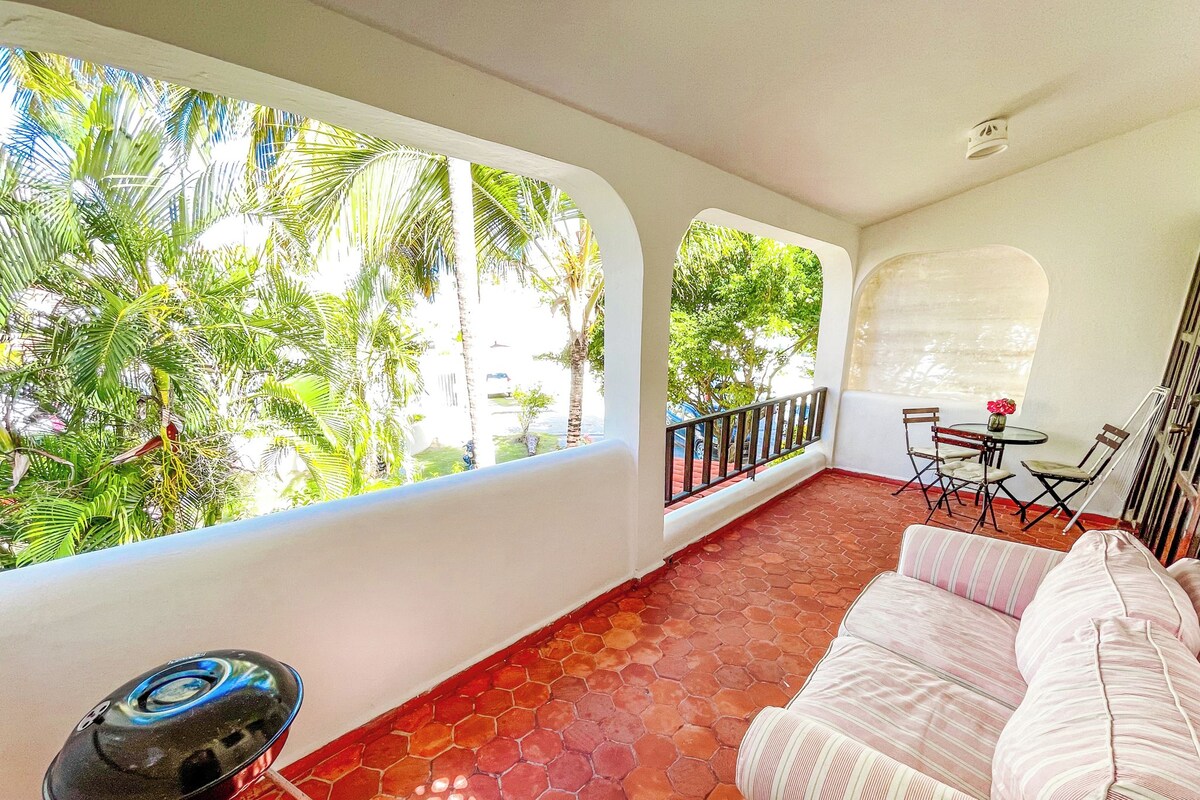 Quarto com piso de telhas vermelhas, sofá branco, mesa pequena com cadeiras e grandes janelas com vista para palmeiras.