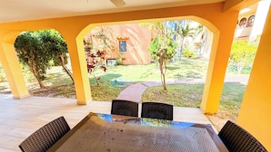 Eine Terrasse im Freien mit Tisch und Stühlen, umgeben von gelben Säulen, mit Blick auf einen Garten mit Bäumen und einem Gebäude im Hintergrund.
