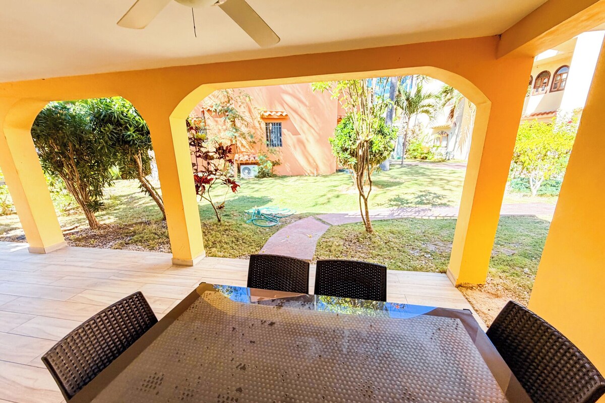 Um pátio externo com mesa e cadeiras, rodeado por pilares amarelos, com vista para um jardim com árvores e um edifício ao fundo.
