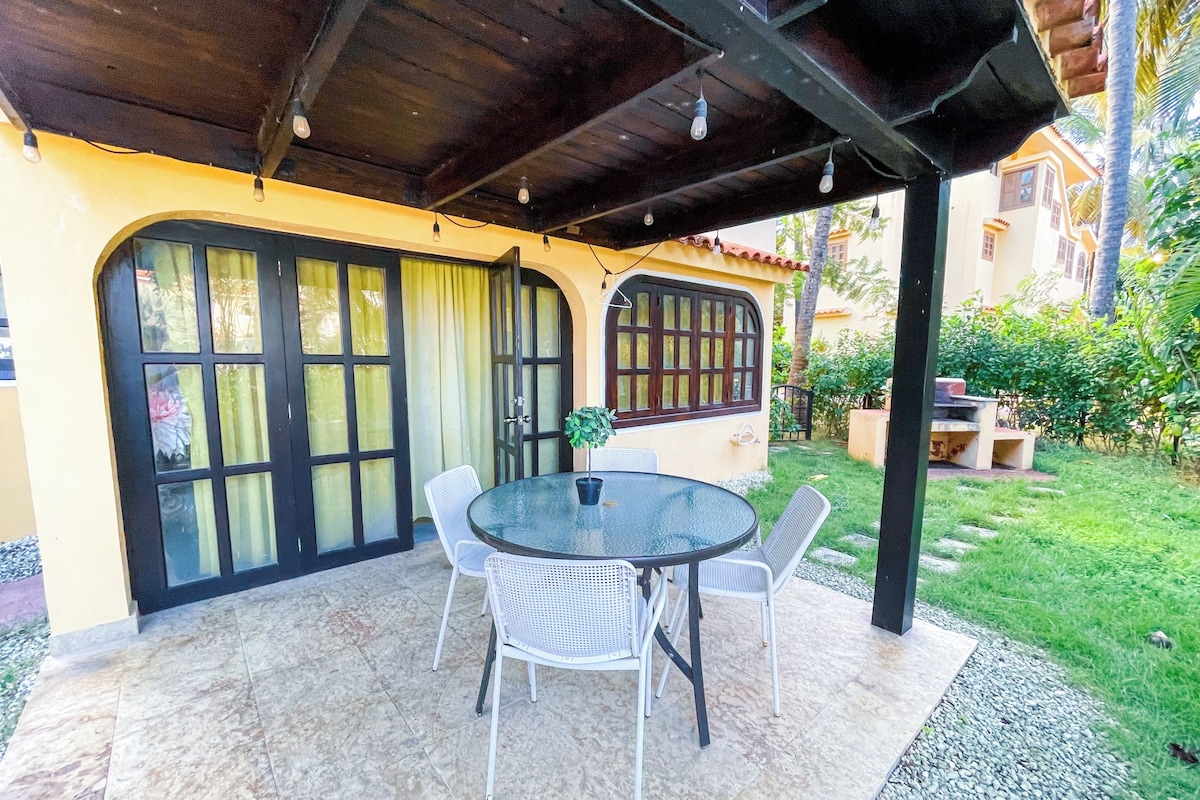 Uma área de estar coberta ao ar livre, com mesa, cadeiras, um vaso de planta e vista para um edifício com grandes janelas e porta.
