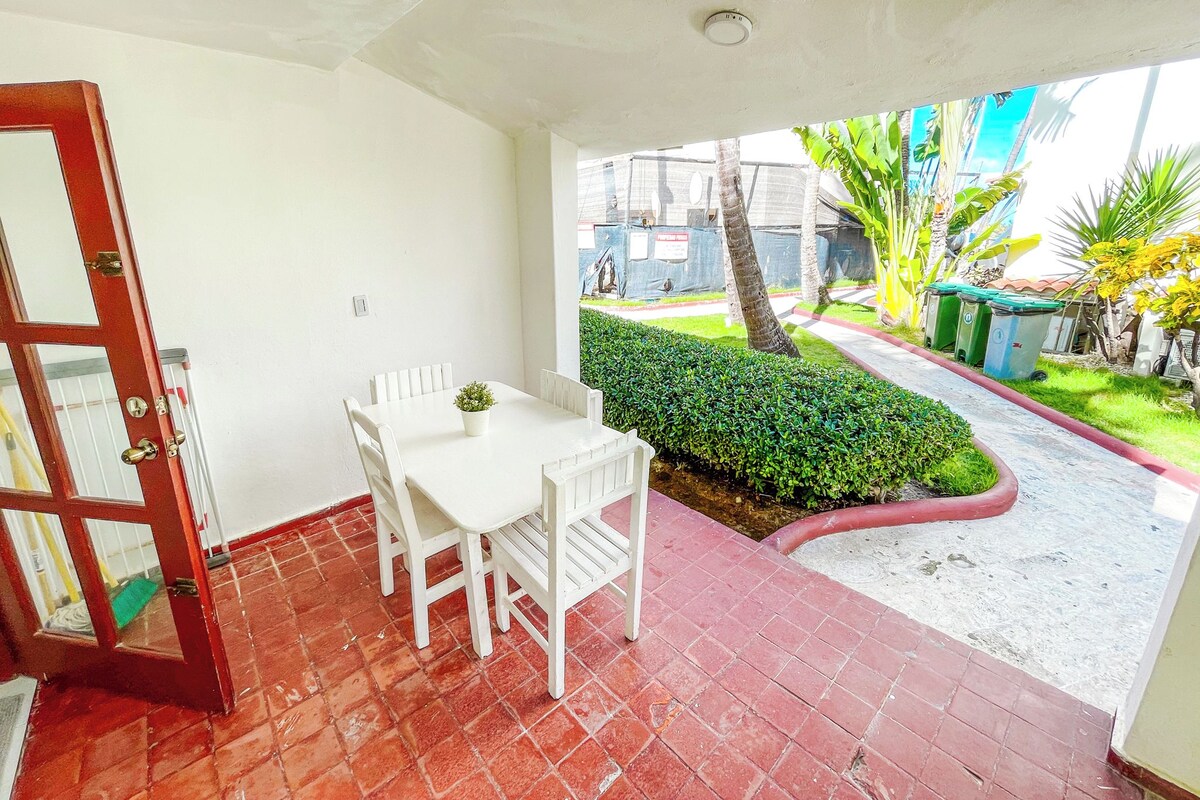 Quarto com mesa e cadeiras brancas, piso de azulejos vermelhos e vista para um jardim com sebes e árvores.