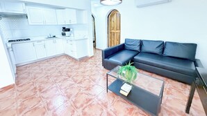 Eine moderne Küche mit weißen Schränken, einem schwarzen Herd und einer Kaffeemaschine. Es gibt ein schwarzes Ledersofa und einen Glastisch mit einer Pflanze.