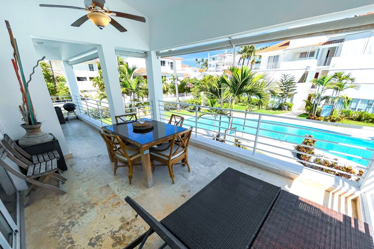 Varanda com mesa e cadeiras, vista para uma piscina e edifícios com vegetação.