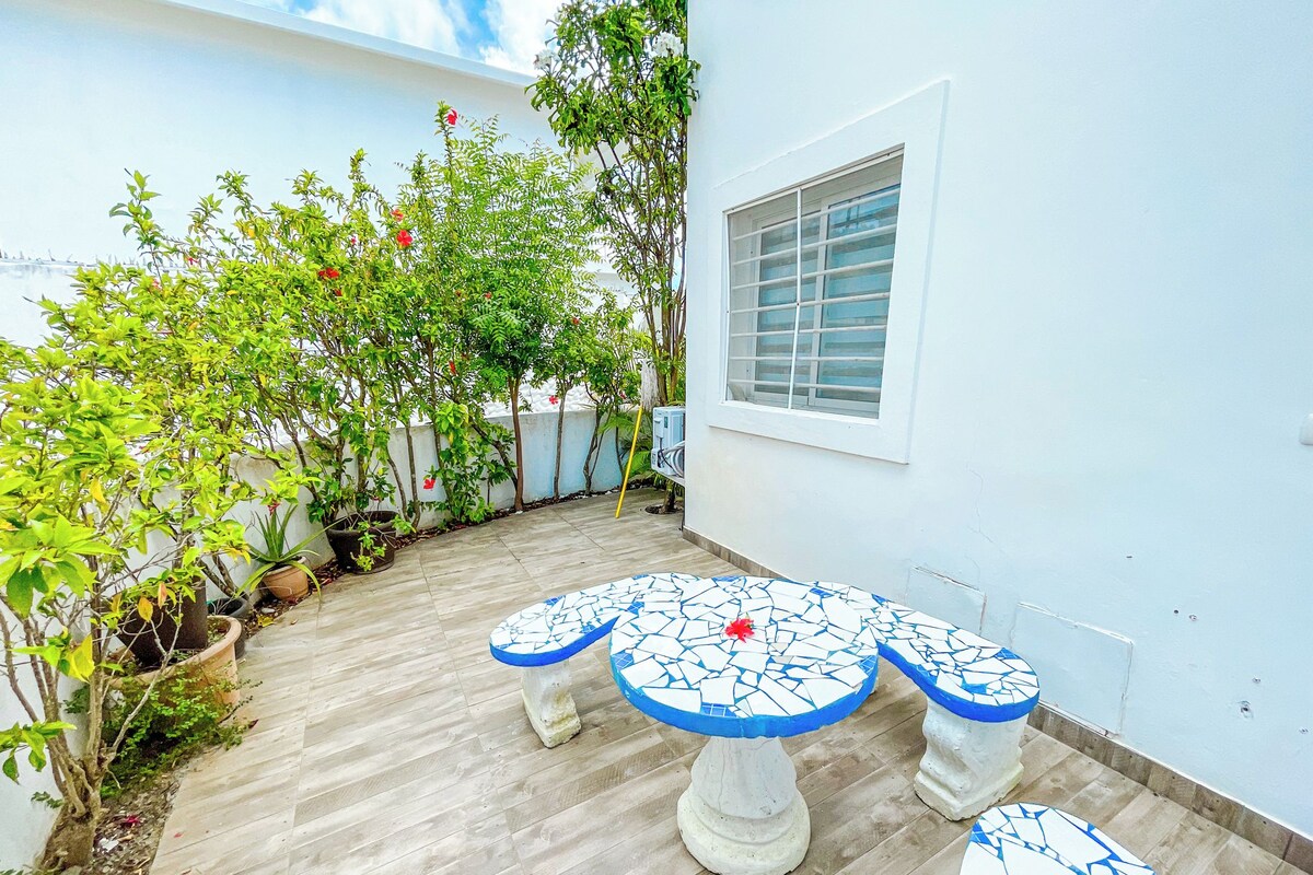 Um pátio com mesa e cadeiras de azulejos, rodeado por plantas em vasos e uma parede branca.