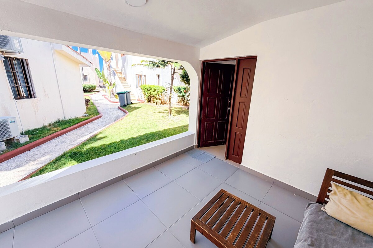Quarto com piso de azulejos, um banco de madeira, uma cama com travesseiro e uma porta que dá para área externa com vista para um edifício e vegetação.