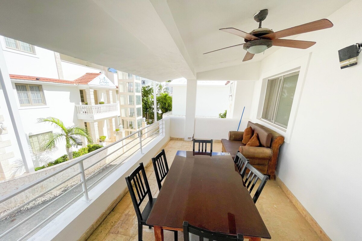 Varanda com mesa e cadeiras de jantar, ventilador de teto e vista para edifícios vizinhos.