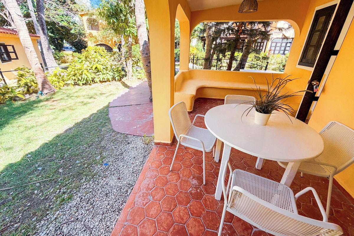 Pátio externo com mesa redonda e cadeiras, cercado por um muro amarelo e vista para o verde.