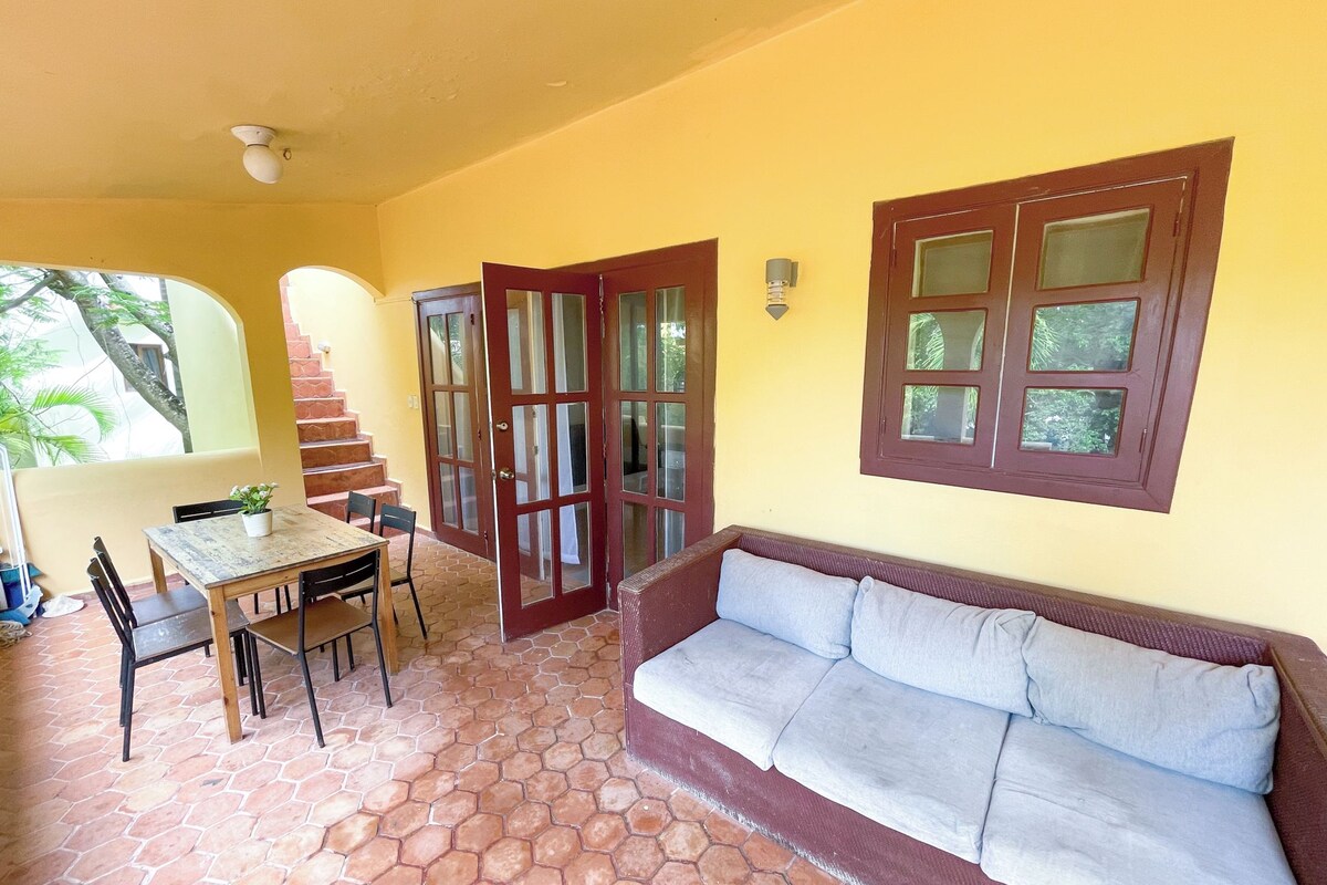Uma varanda coberta com mesa e cadeiras de madeira, um sofá e uma escadaria para um andar superior.