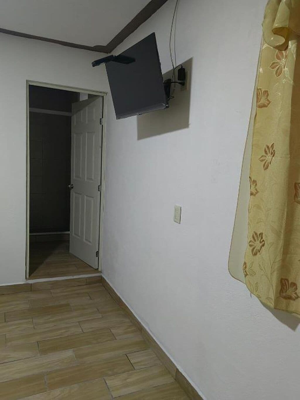 A room with a wooden floor, a closed door, a mounted flat-screen TV, and a curtain with a floral pattern.