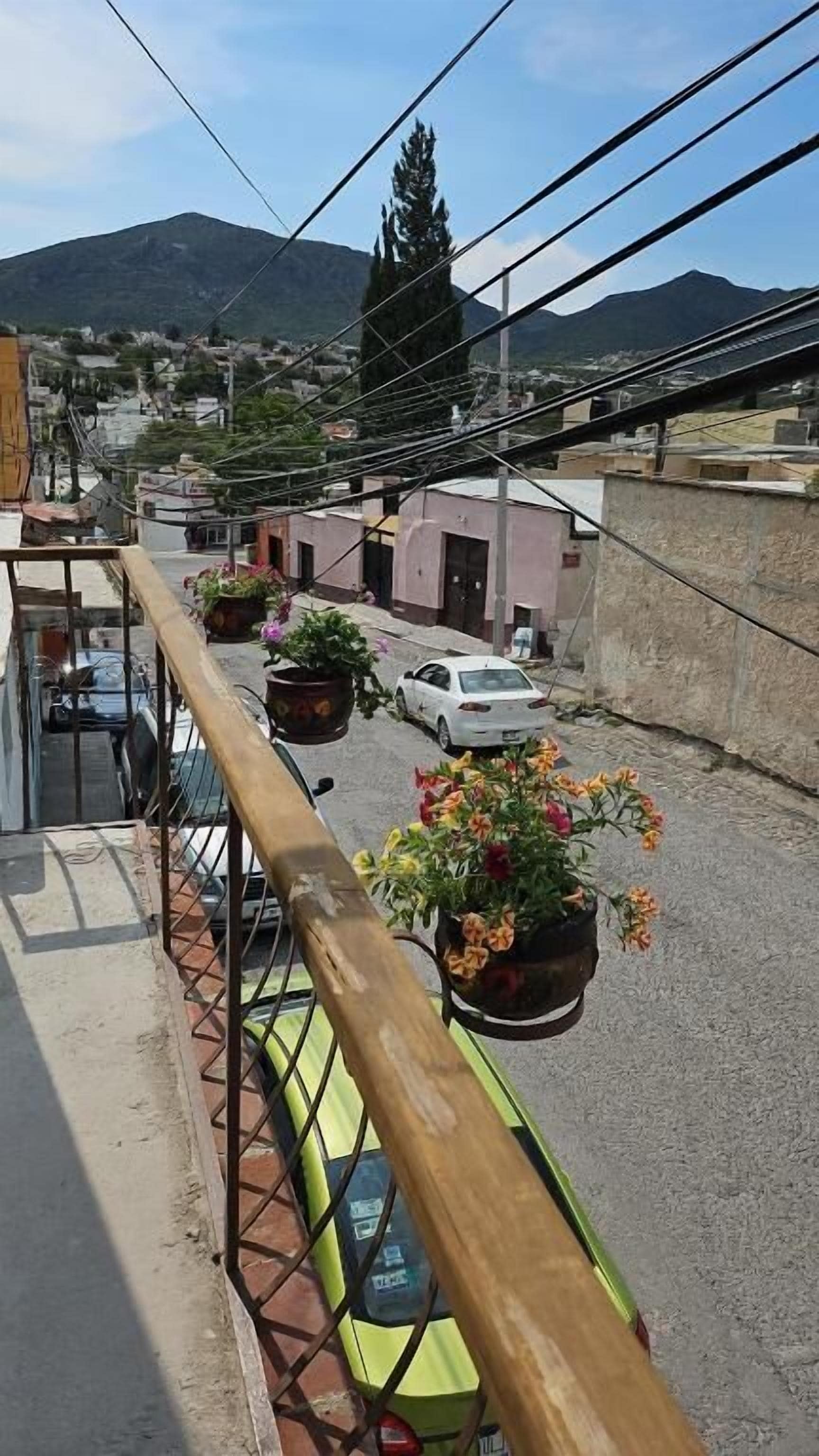 Un balcón con baranda de madera que da a una calle con coches estacionados y edificios, con montañas al fondo.