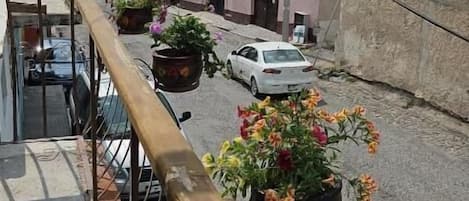 A balcony with a wooden railing overlooking a street with parked cars and buildings, with mountains in the background.