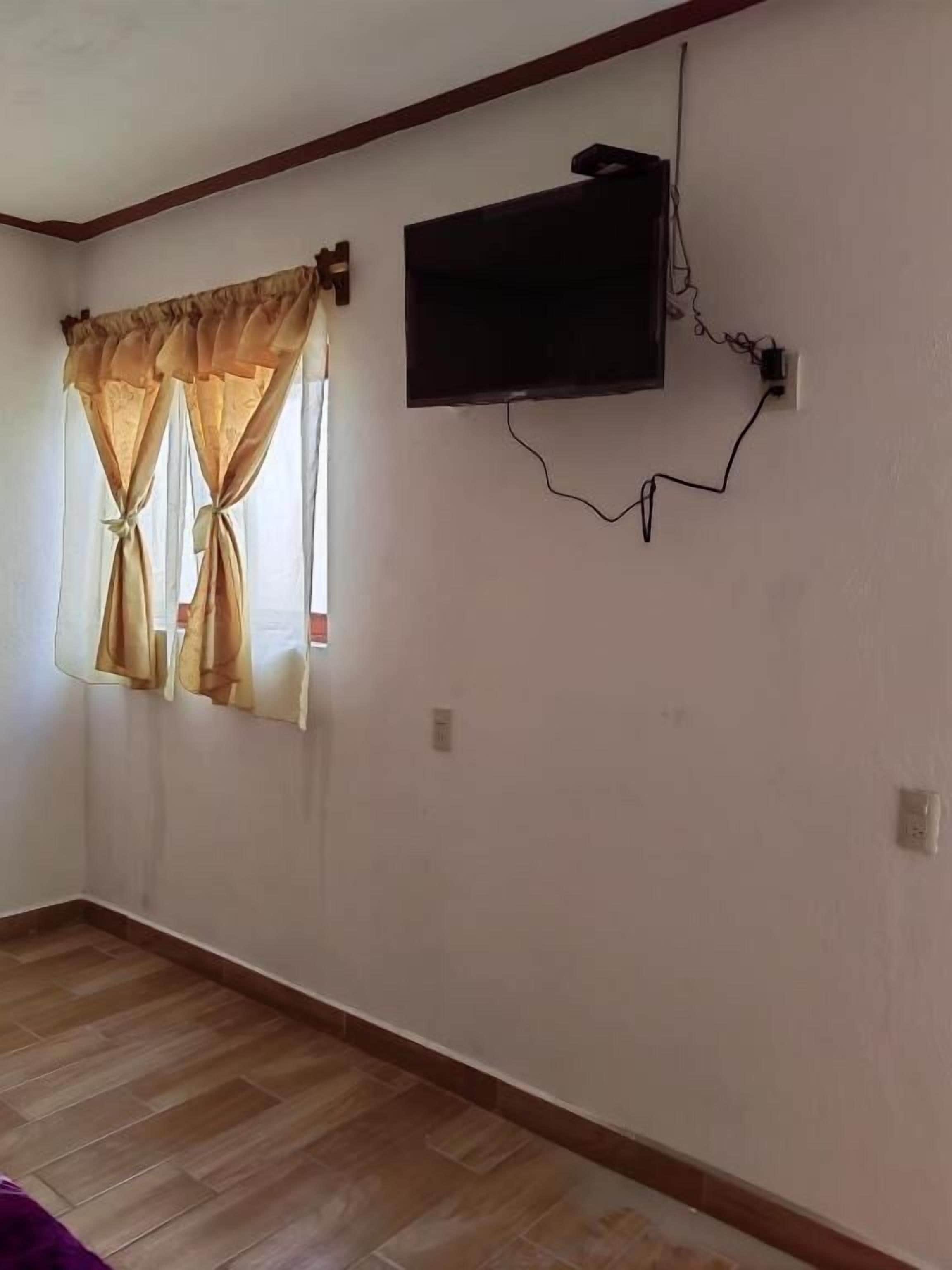 Una habitación con una televisión de pantalla plana montada en la pared, una ventana con cortinas y suelo de madera.